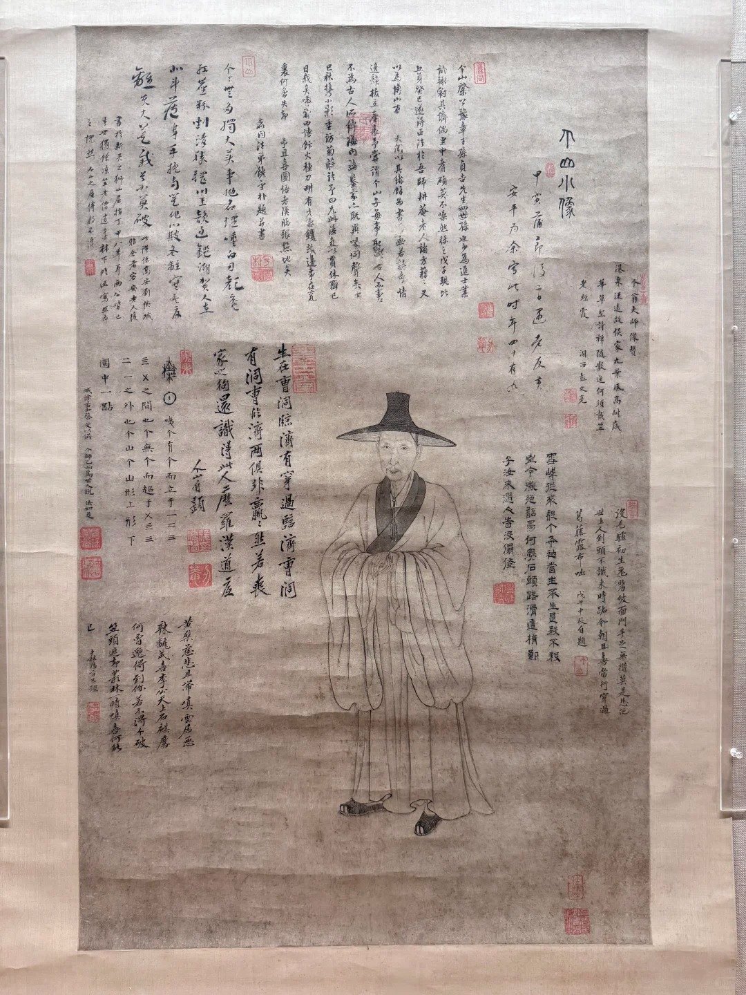 A hanging scroll portrait in ink on paper: a thin, elderly man in a wide dark hat and loose monk's robes stands facing the viewer, drawn almost entirely in fine outline with no shading. The surrounding paper is covered with dense blocks of brushed inscription in several different scripts and stamped with more than a dozen red seals.