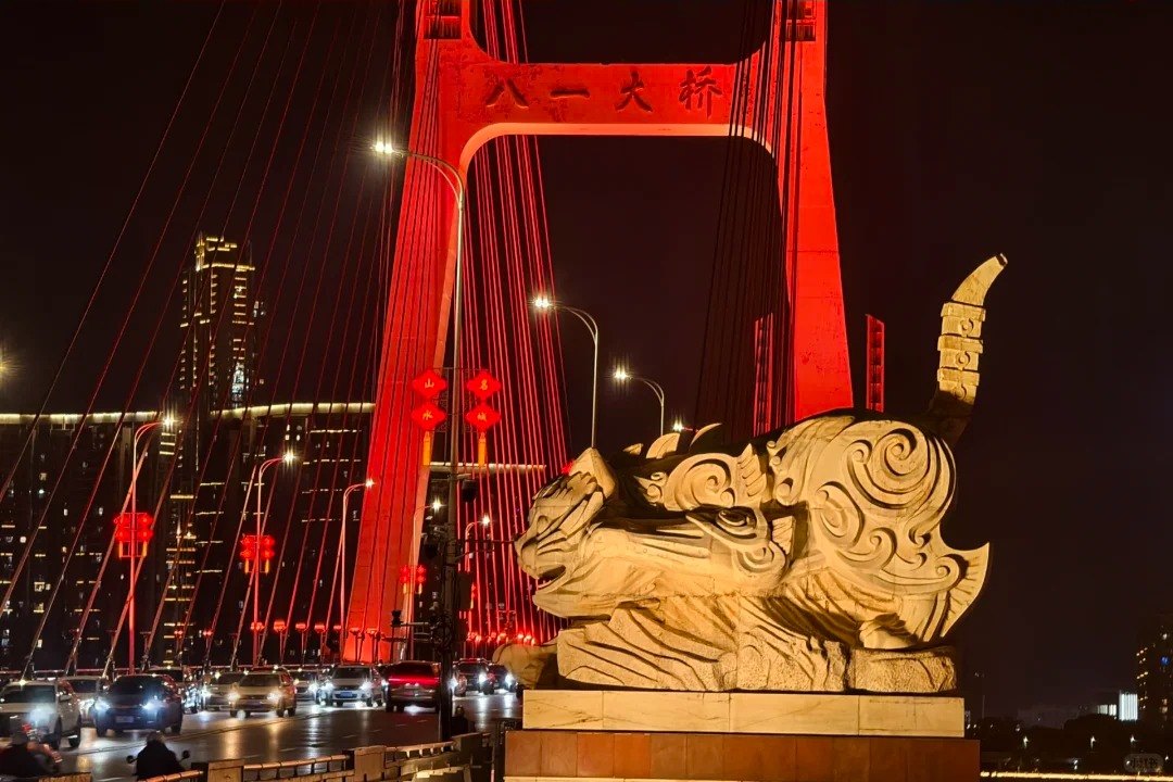 The second Bayi Bridge cat sculpture at night, crouched low along its plinth with the head turned down and the banded tail curling up behind, seen from the rear against the red-lit fan of the bridge stay cables