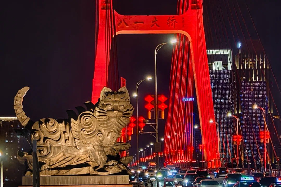 One of the two giant stone cats at the east end of Bayi Bridge at night, sitting upright on its plinth with a banded tail raised behind it and a forepaw reaching over the plinth edge, its flank carved in scrolling cloud motifs, a red-floodlit bridge pylon and a line of red lanterns behind