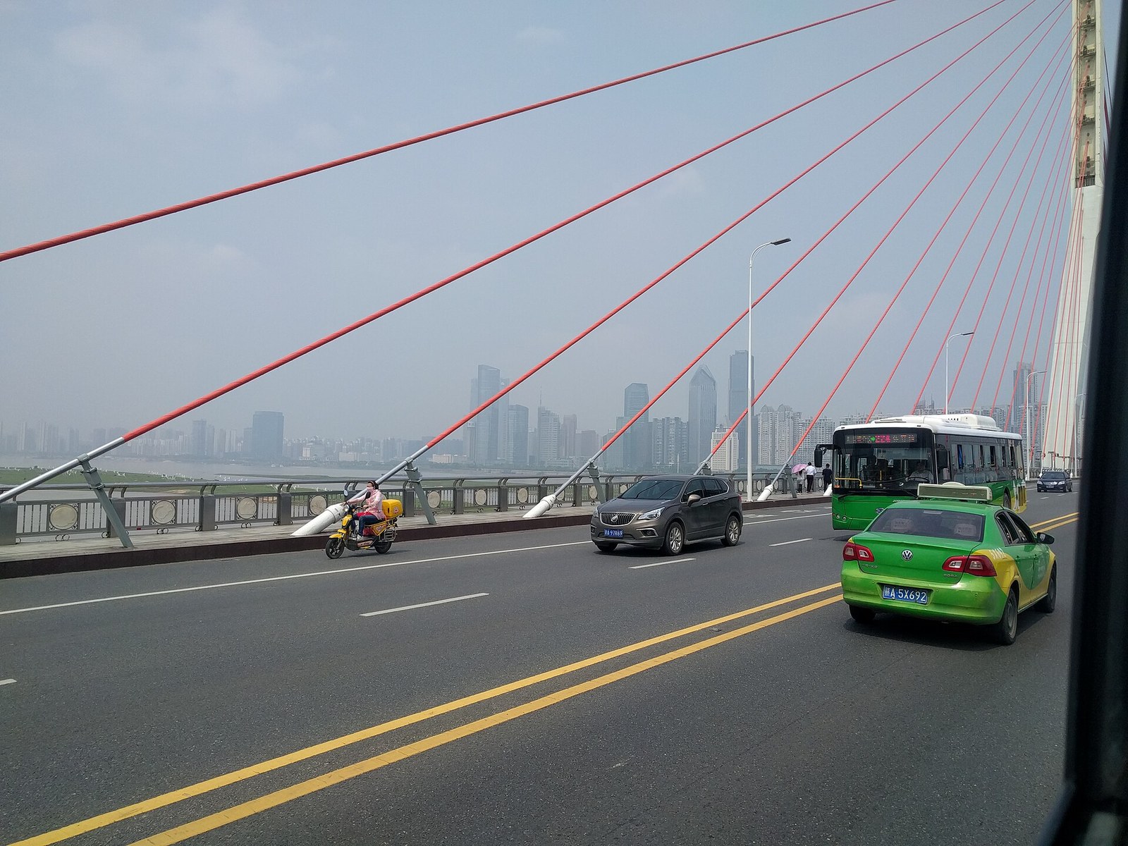 On the deck of Bayi Bridge in Nanchang: red stay cables raking down to the deck, a separated side lane with a scooter, parapet panels carrying carved roundels, and the Honggutan skyline hazy ahead