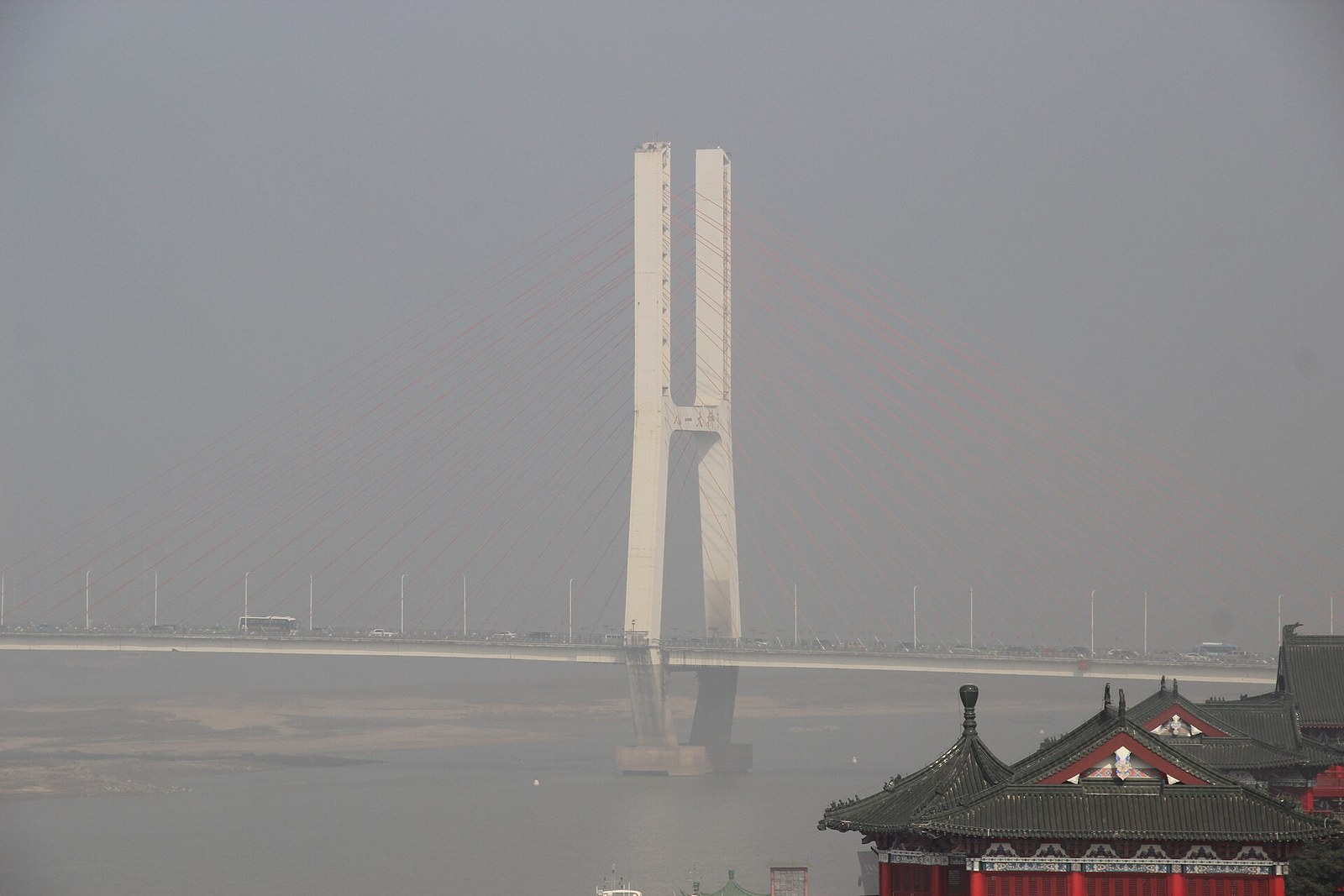 Bayi Bridge across the Gan River in Nanchang seen from Tengwang Pavilion, showing one H-shaped white pylon carrying fans of red stay cables above the traffic deck on a hazy day