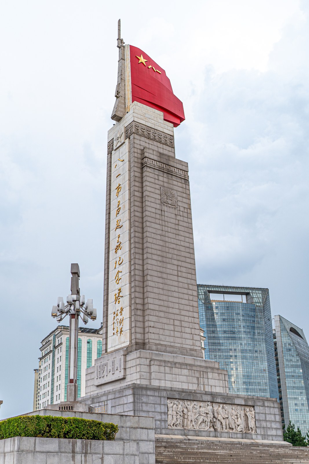 The August 1st Uprising Monument in Nanchang by day, showing the red army-flag sculpture and stone rifle at the top and gilded calligraphy down the granite shaft