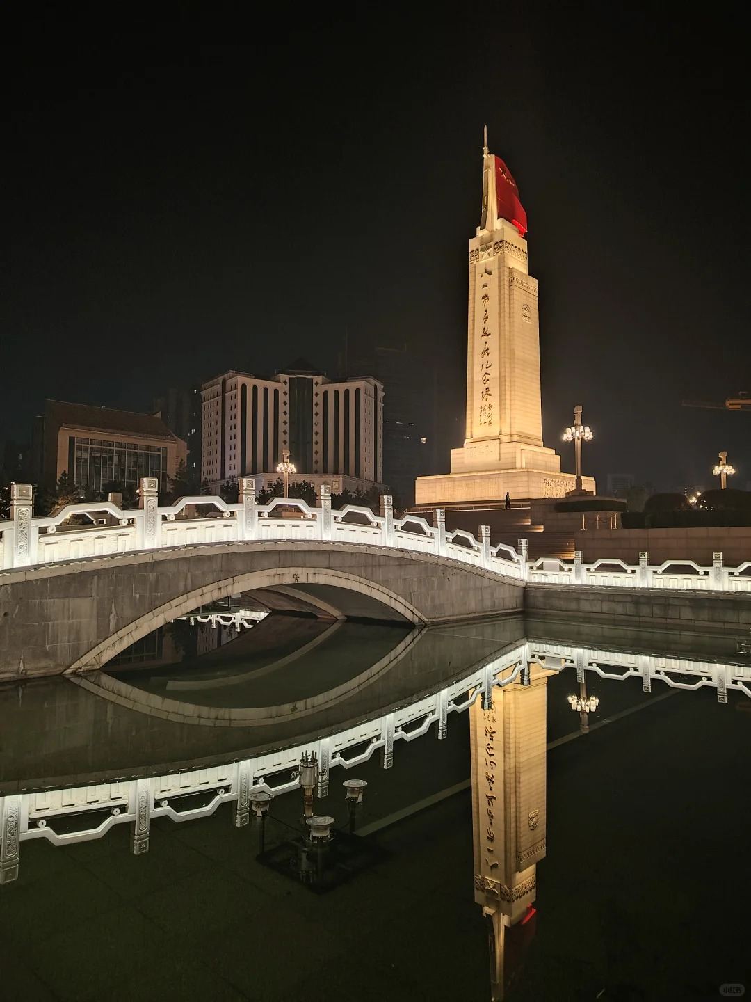 The August 1st Uprising Monument lit warm at night, reflected in the Golden Water canal beside a white arched stone bridge in Bayi Square, Nanchang