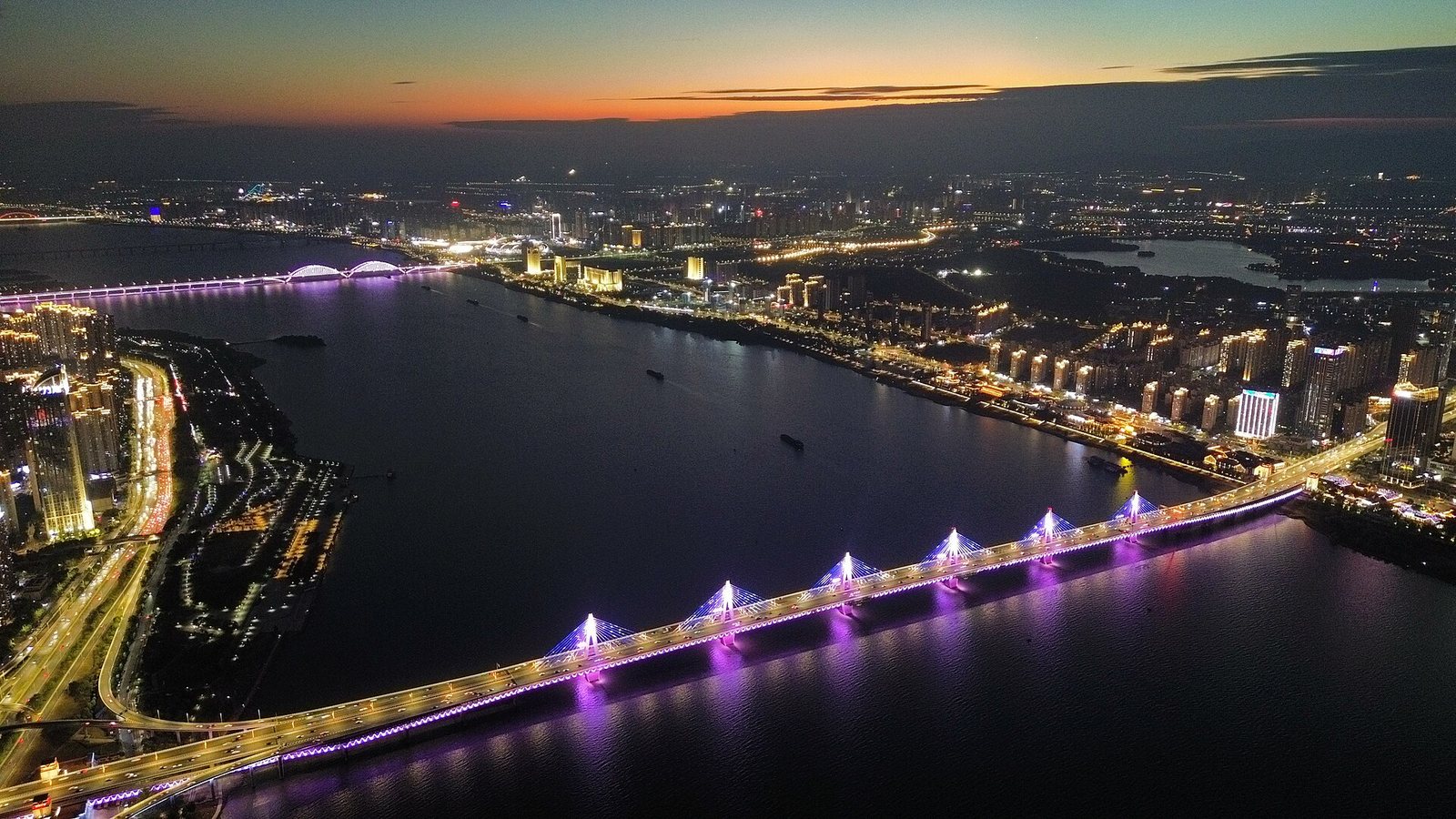 Chaoyang Bridge in Nanchang lit purple and blue at dusk, seen from the air: six slim pylons with a single central plane of stay cables crossing the Gan River, city lights on both banks