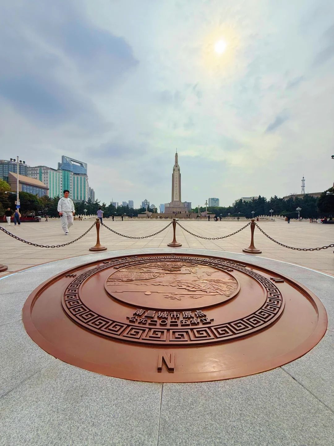 The bronze City Origin Point marker set into the granite of Bayi Square, Nanchang — three concentric rings around a relief map of the city, with the August 1st Uprising Monument rising behind on the square's axis
