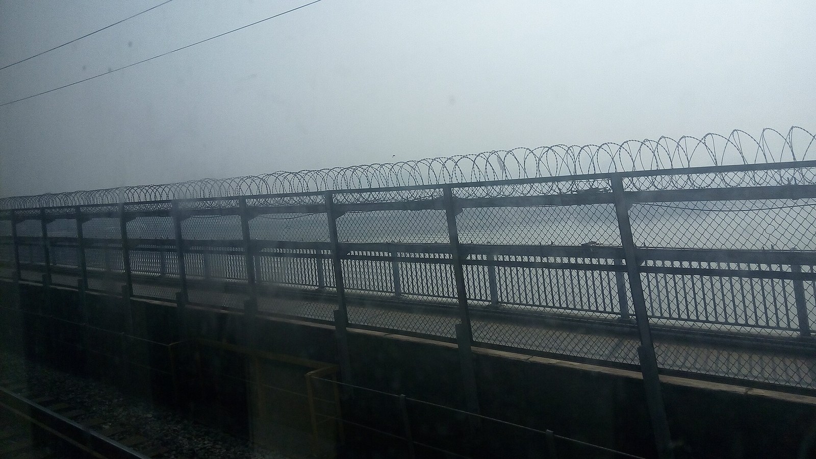 The 1963 Gan River Bridge in Nanchang from a train window: ballasted railway track in the foreground, a chain-link fence topped with razor wire, and the road carriageway immediately beyond it over misty water
