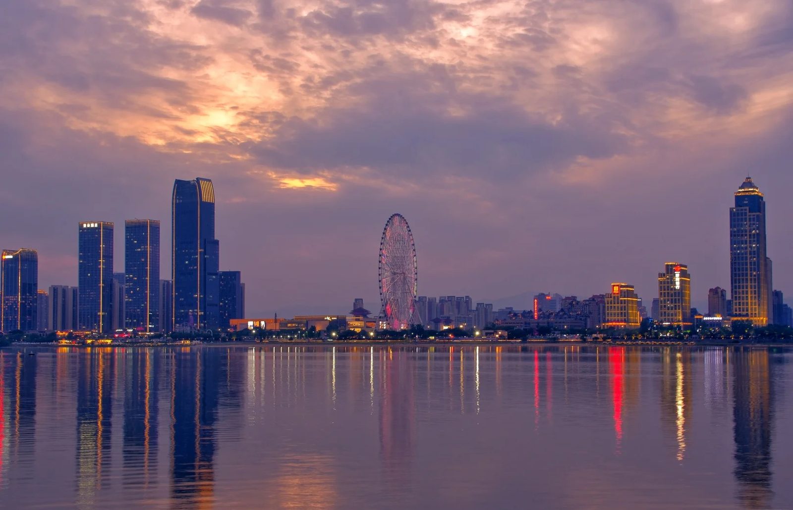 The Honggutan skyline in Nanchang at dusk under a violet and orange sky, its lit towers and the Star of Nanchang Ferris wheel reflected in the still water of the Gan River