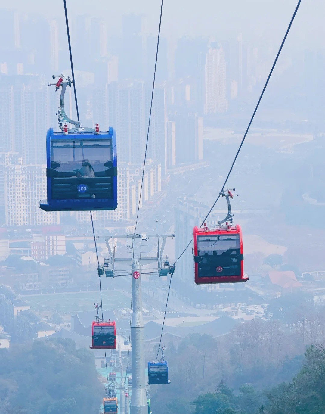 Blue and red gondola cabins on the Meiling cableway above Nanchang, hanging from cables over a support tower with hazy tower blocks and woodland far below