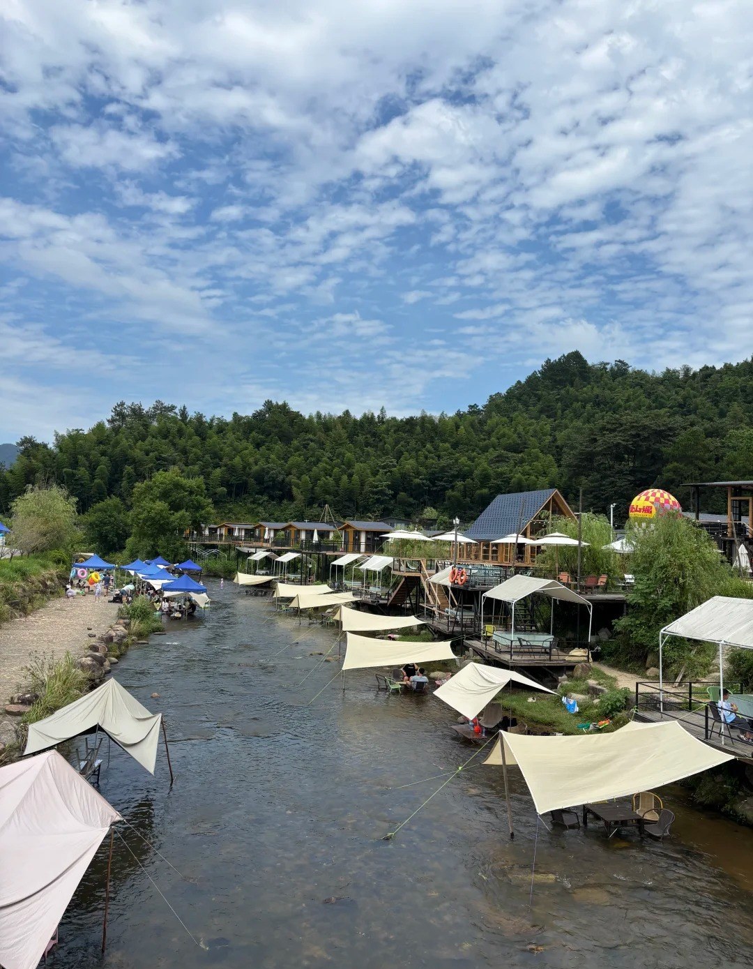 Cream canopies rigged over a shallow clear stream at Meiling, Nanchang, with tables and chairs standing in the water, timber cabins and decks along the right bank and blue tents and people paddling on the left