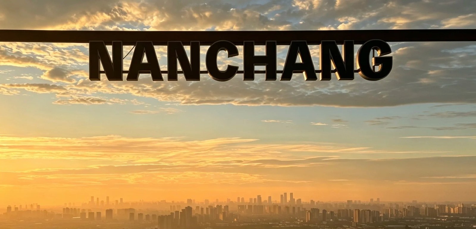 Sunrise from the Panlong Peak viewing deck at Meiling, Nanchang: large black three-dimensional NANCHANG letters on the deck rail silhouetted against an orange sky, with the city skyline hazy on the horizon