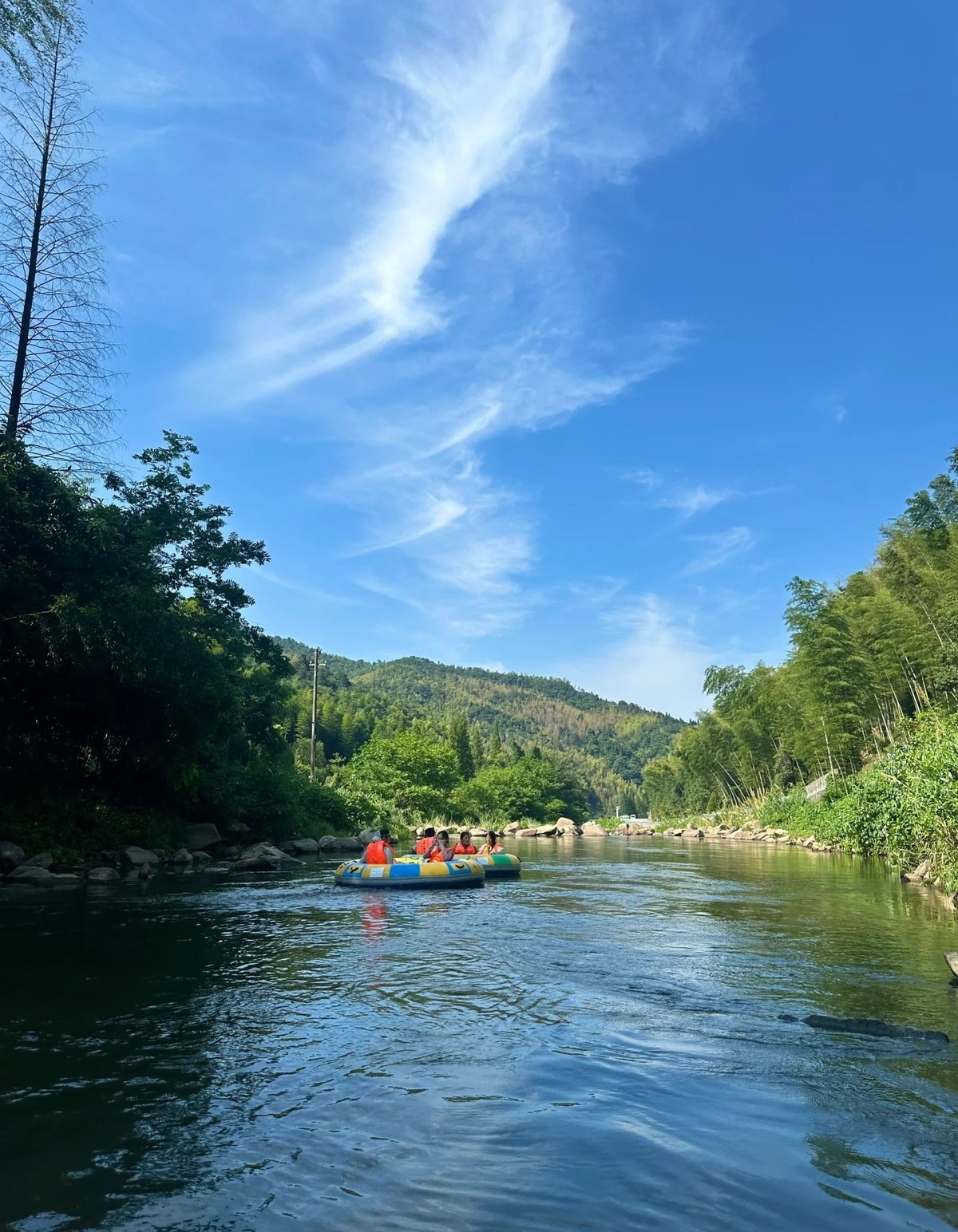A yellow and blue inflatable raft carrying passengers in orange life jackets down a calm green stream at Meiling, Nanchang, with bamboo on the banks and forested hills behind
