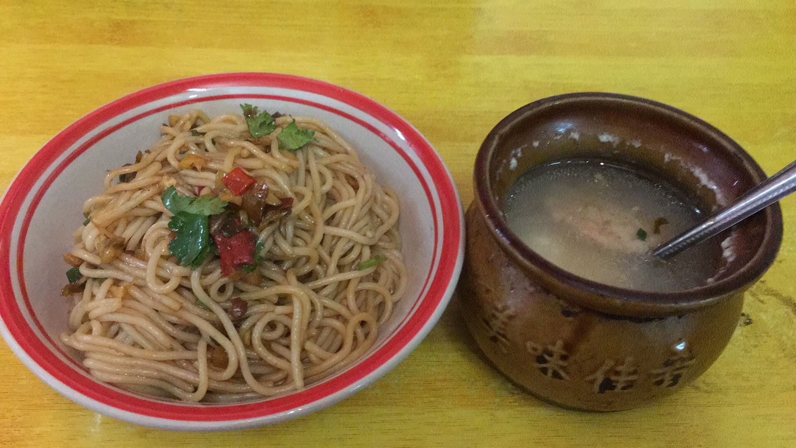 A bowl of Nanchang banfen, round rice noodles tossed with chilli, pickles, peanuts and scallion, beside a small clay pot of soup on a table