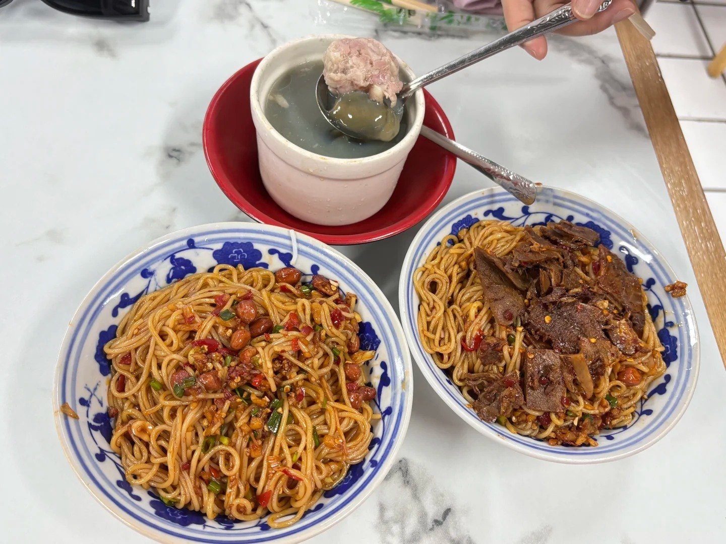 Two blue-and-white bowls of chilli-tossed noodles beside a white clay pot of soup, with a pale meat patty lifted clear of the broth on a spoon.