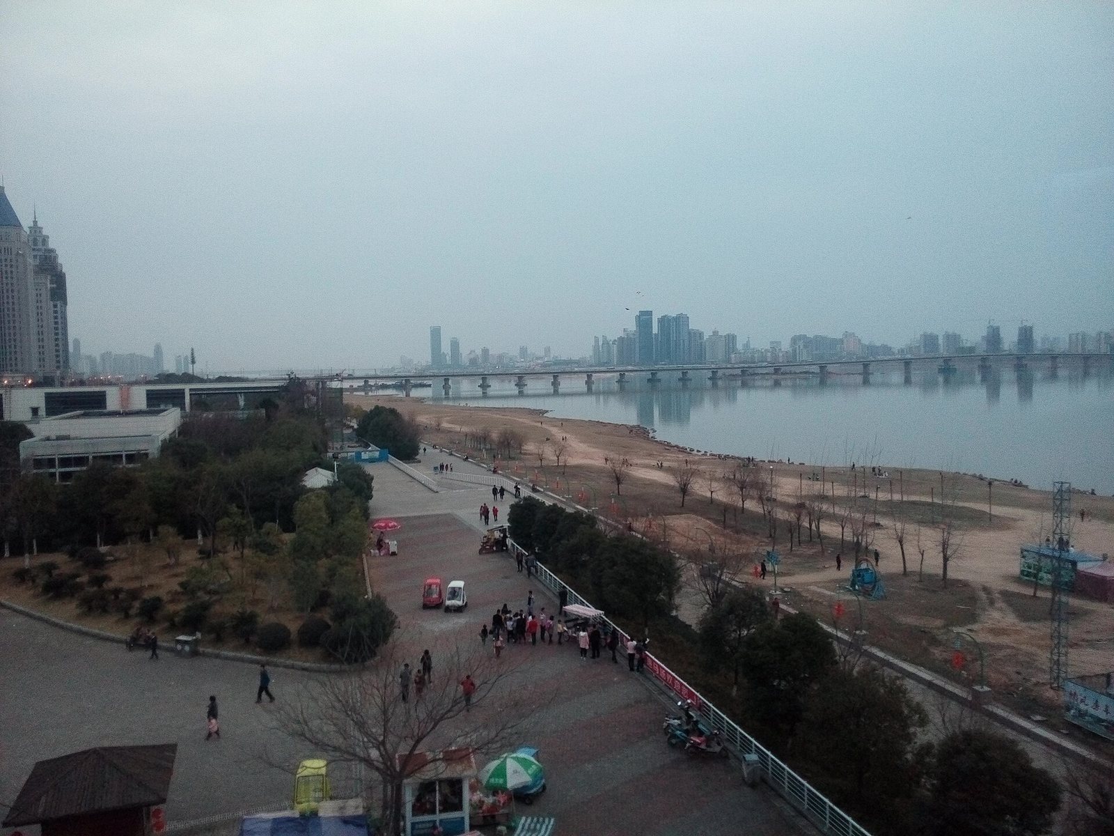 Nanchang Bridge crossing the Gan River, a long low multi-span beam bridge seen from an elevated riverside park where people are walking on the embankment above an exposed sandy bank