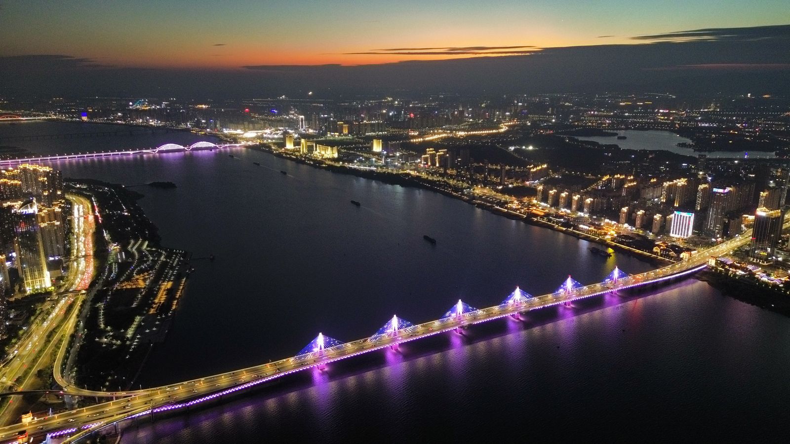 Aerial view of the Gan River in Nanchang at dusk, a lit bridge crossing the water and the Honggutan riverfront glowing under an orange sky