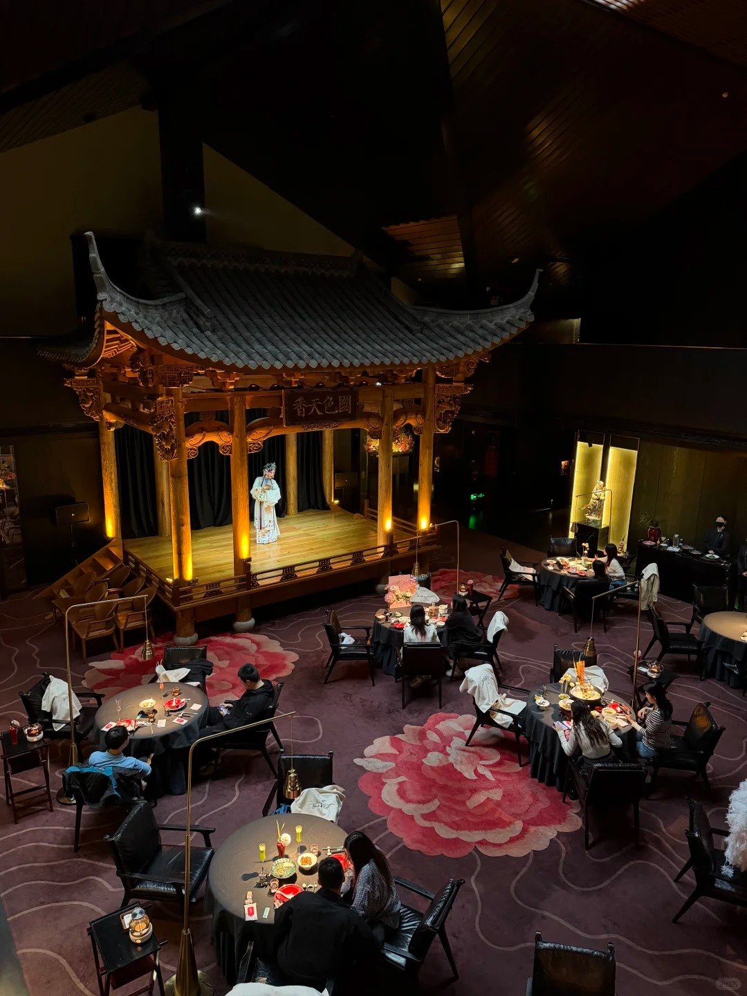 Looking down on a dark restaurant hall where round dining tables surround a lit wooden opera stage, with a costumed performer on it and a peony-patterned carpet across the floor.