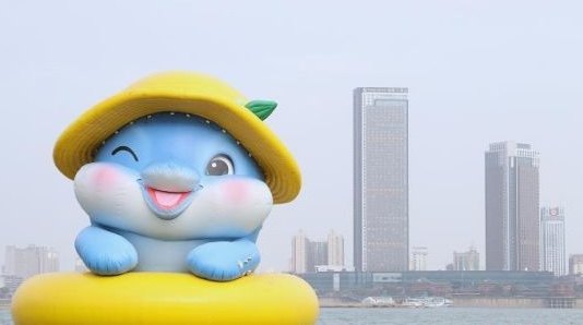 Tun Bao, a giant blue inflatable Yangtze finless porpoise mascot in a yellow sun hat and yellow rubber ring, floating on the Gan River at Qiushui Square in Nanchang with high-rise towers on the far bank behind it