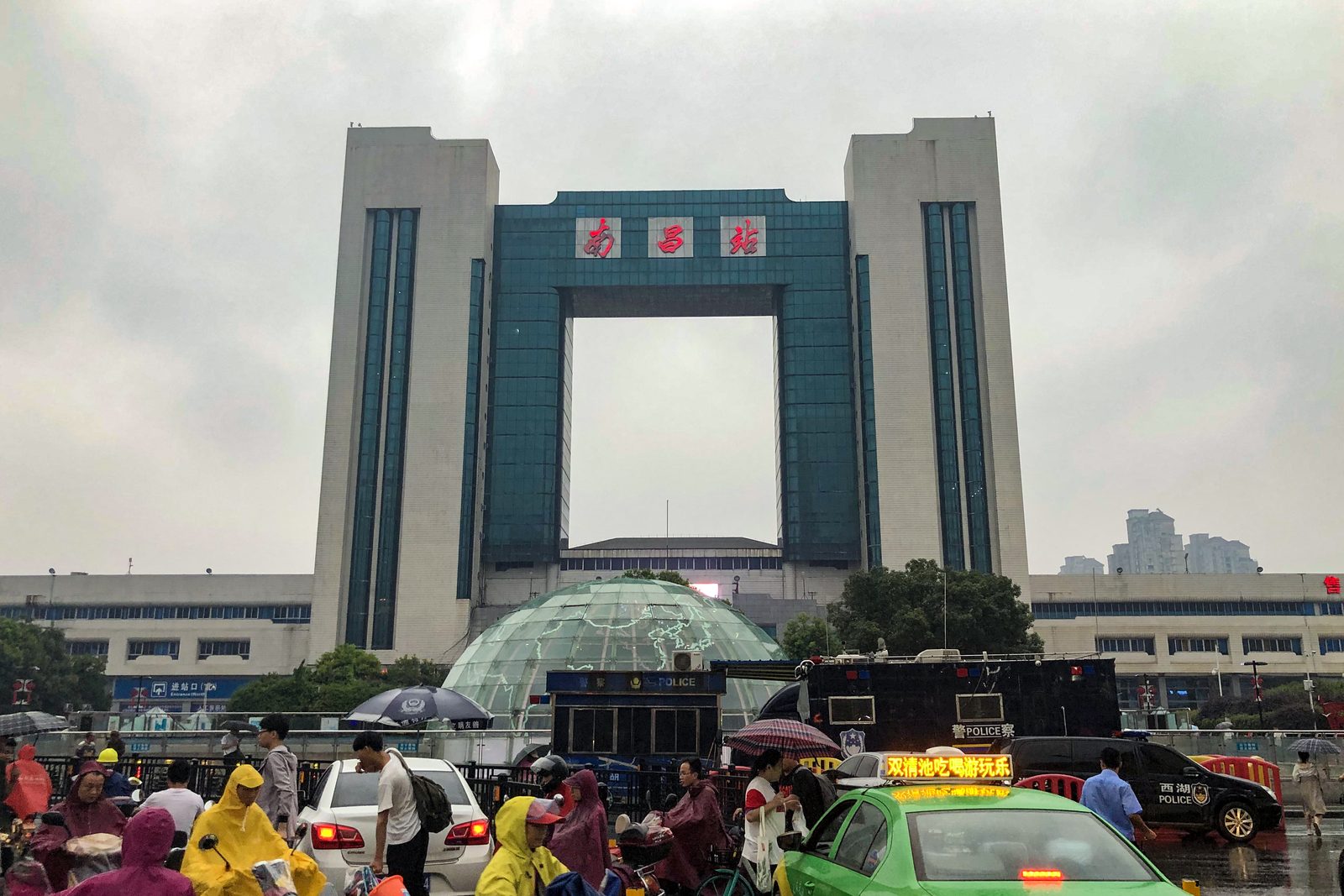 The gateway-arch main building of Nanchang Railway Station with its red name sign and a glass-domed entrance, taxis and travellers out front