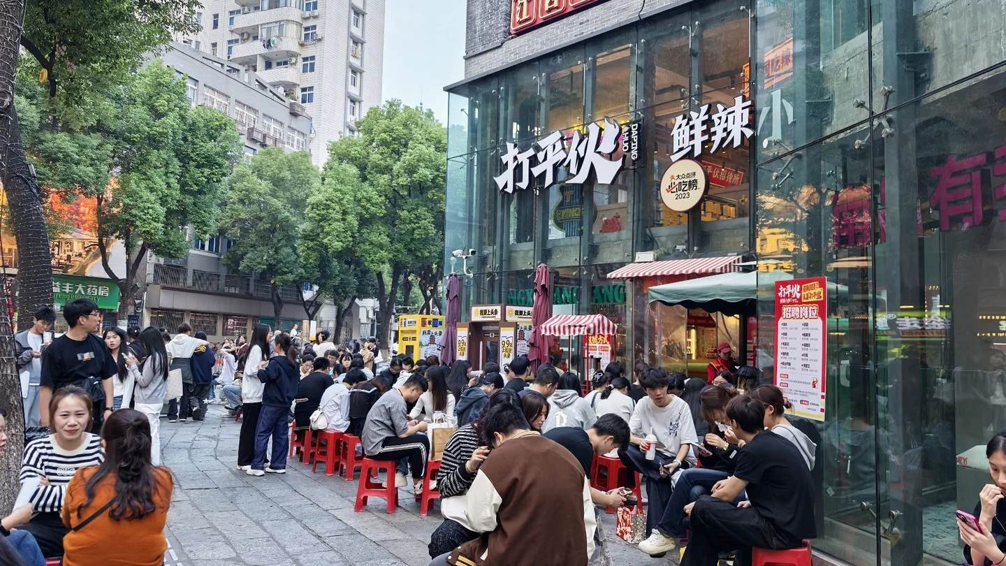 Dozens of people sitting on red plastic stools along a Nanchang pavement outside a spicy stir-fry restaurant, waiting for a table in daylight.
