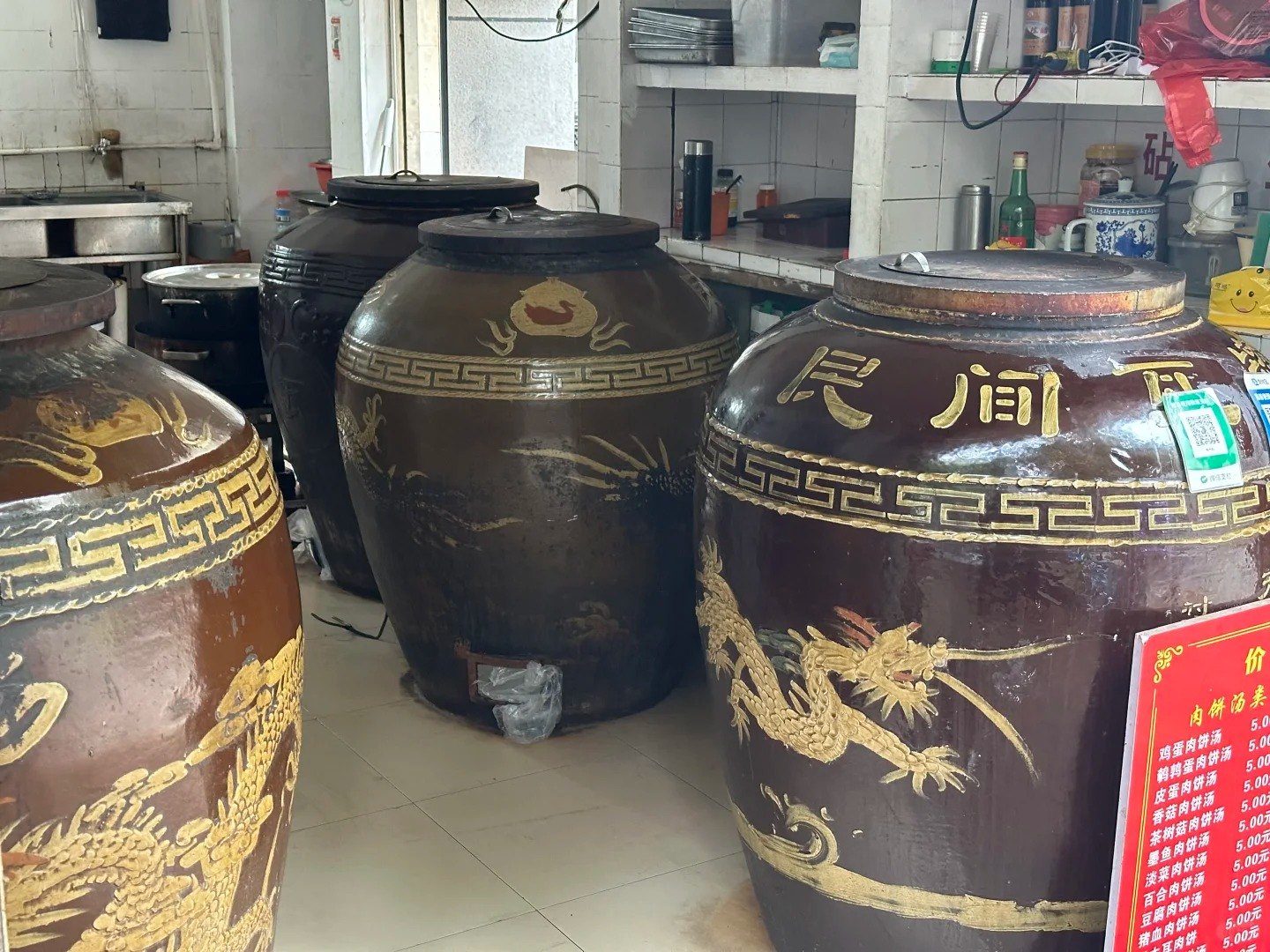 Four large glazed ceramic urns with gold dragon motifs and heavy lids in a Nanchang soup shop, beside a red price board listing meat-patty soups.
