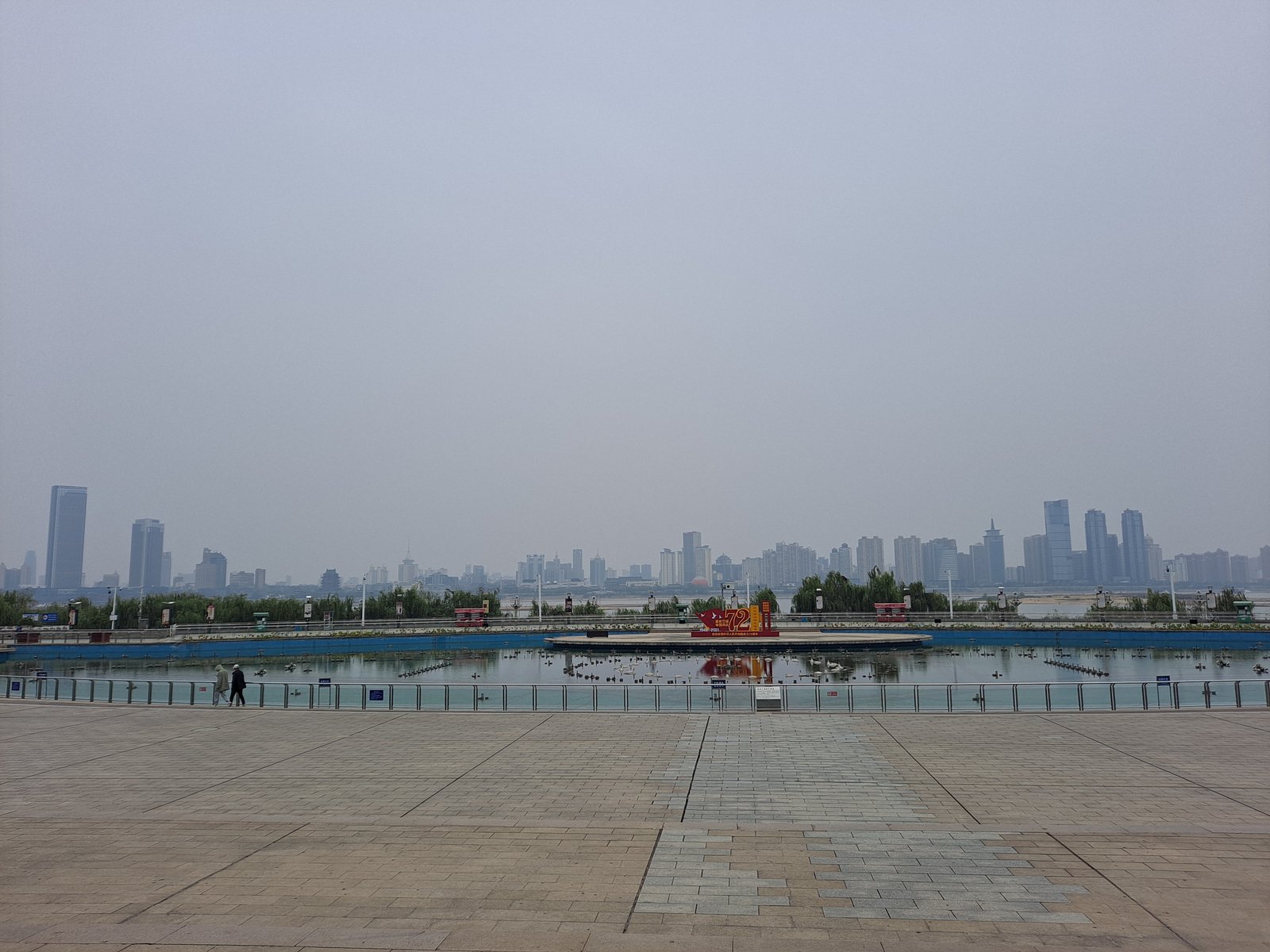 Qiushui Square in Nanchang by day: a vast paved riverside plaza with the fountain platform at rest and the city skyline hazy across the Gan River