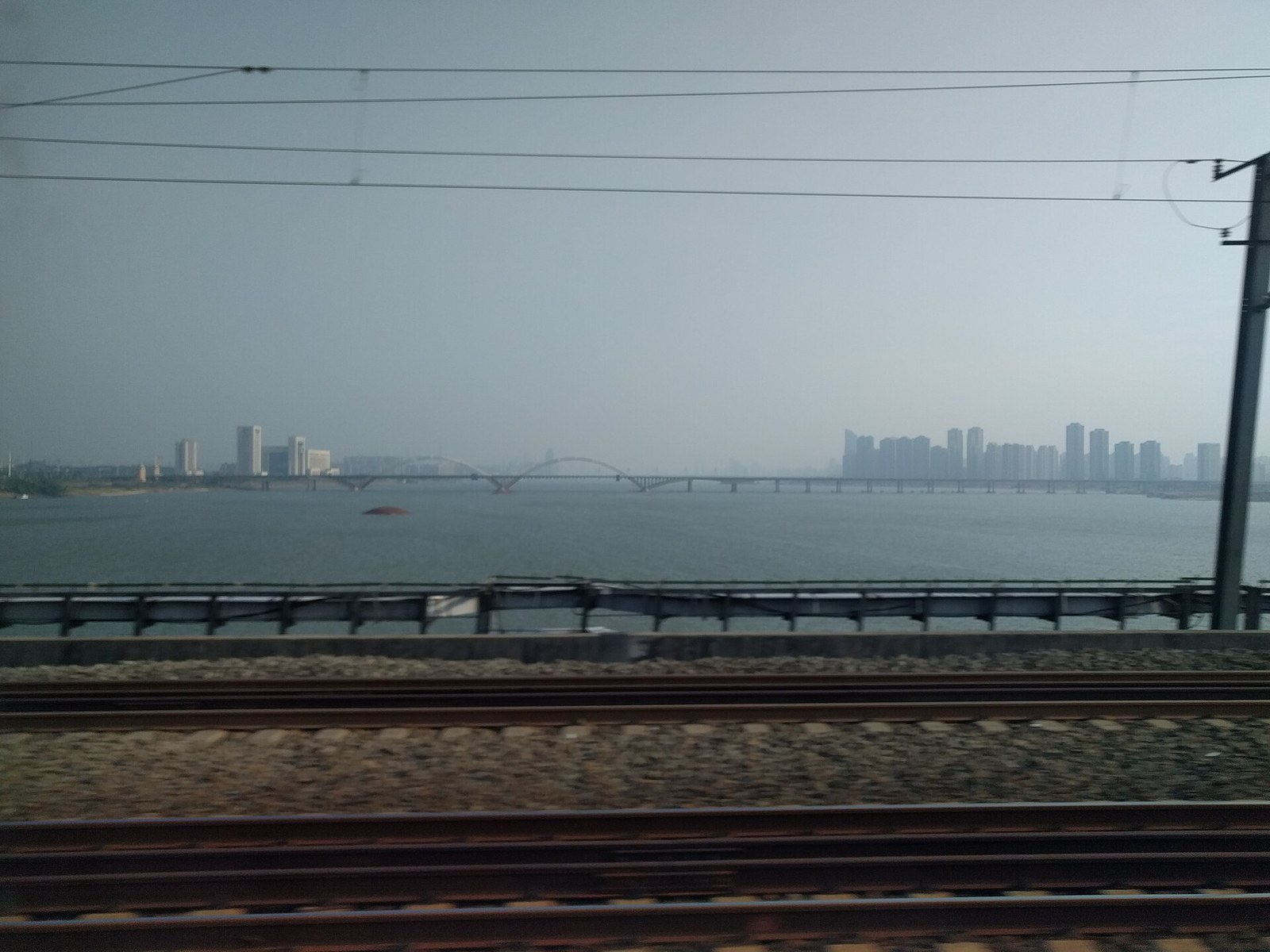 Shengmi Bridge in Nanchang, its twin steel tied arches spanning the wide grey Gan River, photographed from a passing train with railway track in the foreground