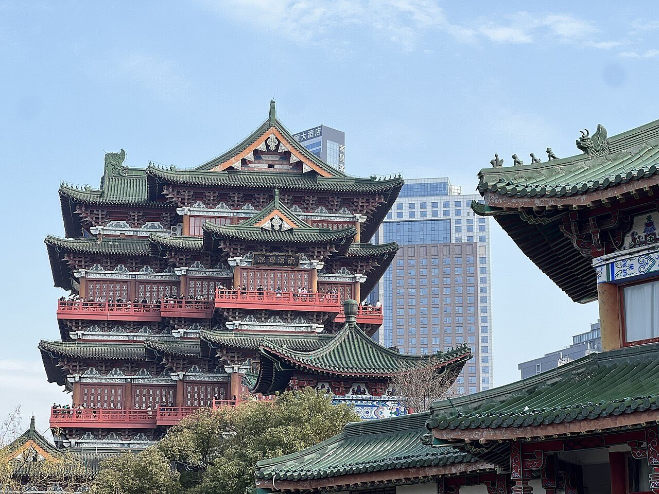 Tengwang Pavilion seen from the side in daylight, its green-glazed eaves and red verandah balustrades stacked in tiers with visitors leaning on the rails, and a wall of modern glass and concrete office towers rising directly behind it.