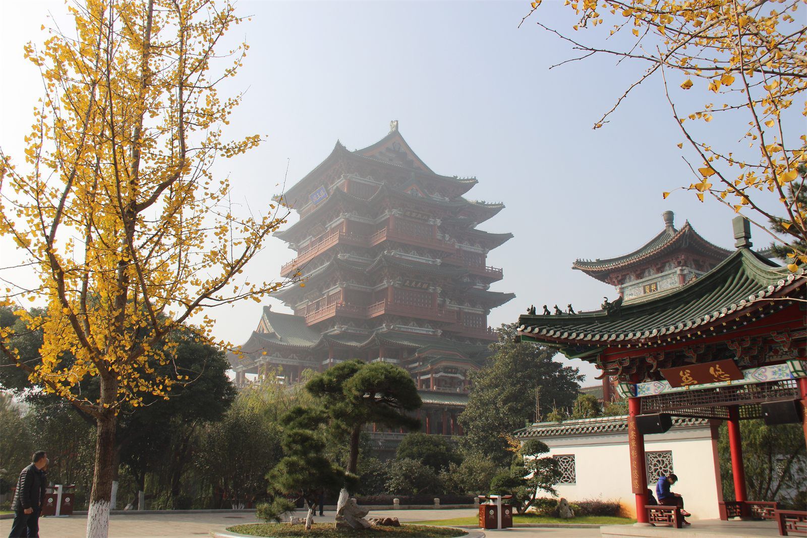 Tengwang Pavilion in Nanchang — a tall, many-storeyed timber-style tower with sweeping jade-green glazed roofs and vermilion pillars, seen across the courtyard through autumn ginkgo.