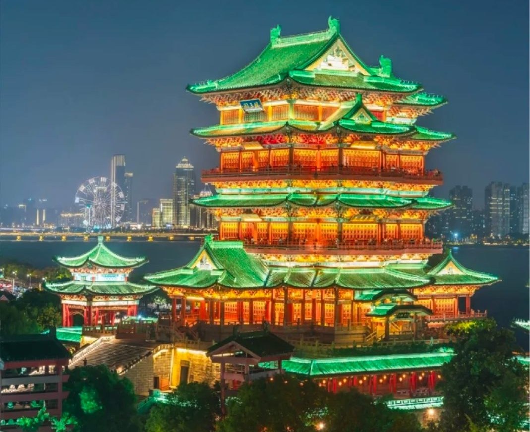 Tengwang Pavilion floodlit after dark, its glazed roofs washed green and the open verandah levels lit warm amber, with the Gan River, a lit bridge, the Star of Nanchang Ferris wheel and the Honggutan skyline behind it.