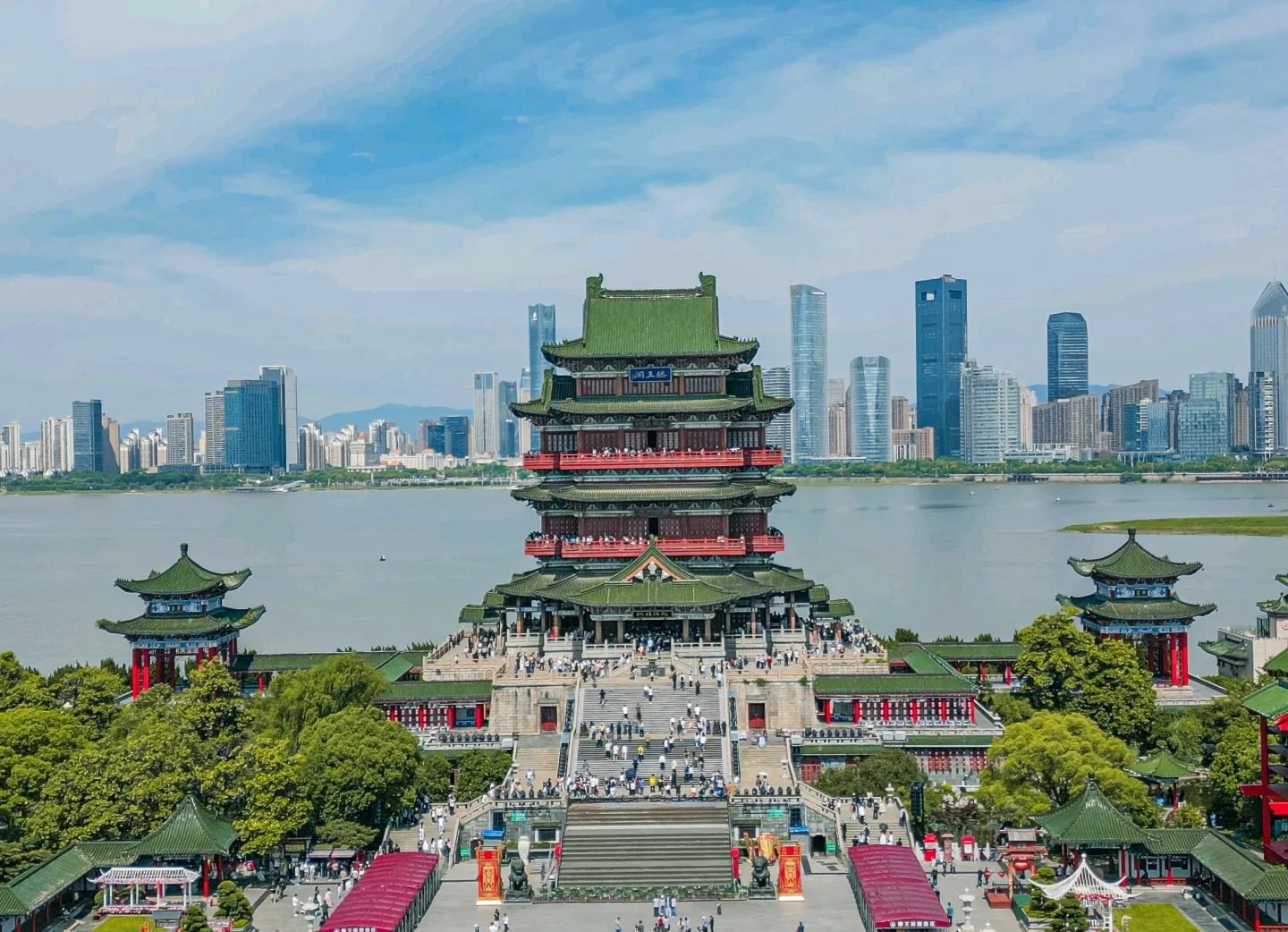 Tengwang Pavilion in Nanchang seen from above on a clear day, its green tiled roofs and red balconies rising above crowded stone steps, with the Gan River and the Honggutan high-rise skyline behind it