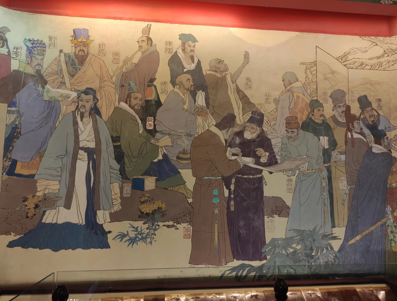 A wide painted mural of some twenty robed historical figures from Jiangxi arranged across a pale landscape, each labelled with a small red cartouche of characters, two of them unrolling a scroll in the centre foreground.
