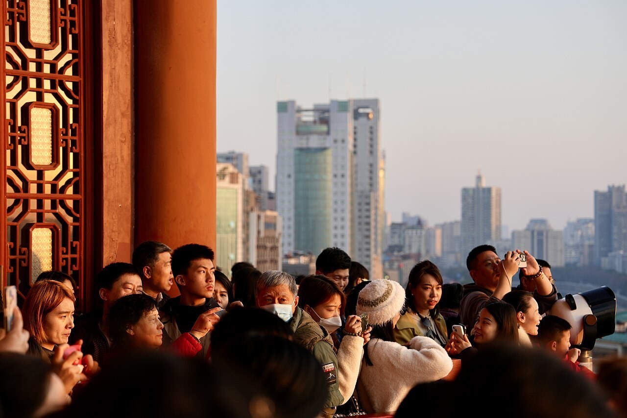 Visitors pressed shoulder to shoulder along the top-gallery railing of Tengwang Pavilion in low golden light, phones raised, a vermilion column and latticework window at the left, the hazy city skyline beyond and a coin-operated telescope at the right.