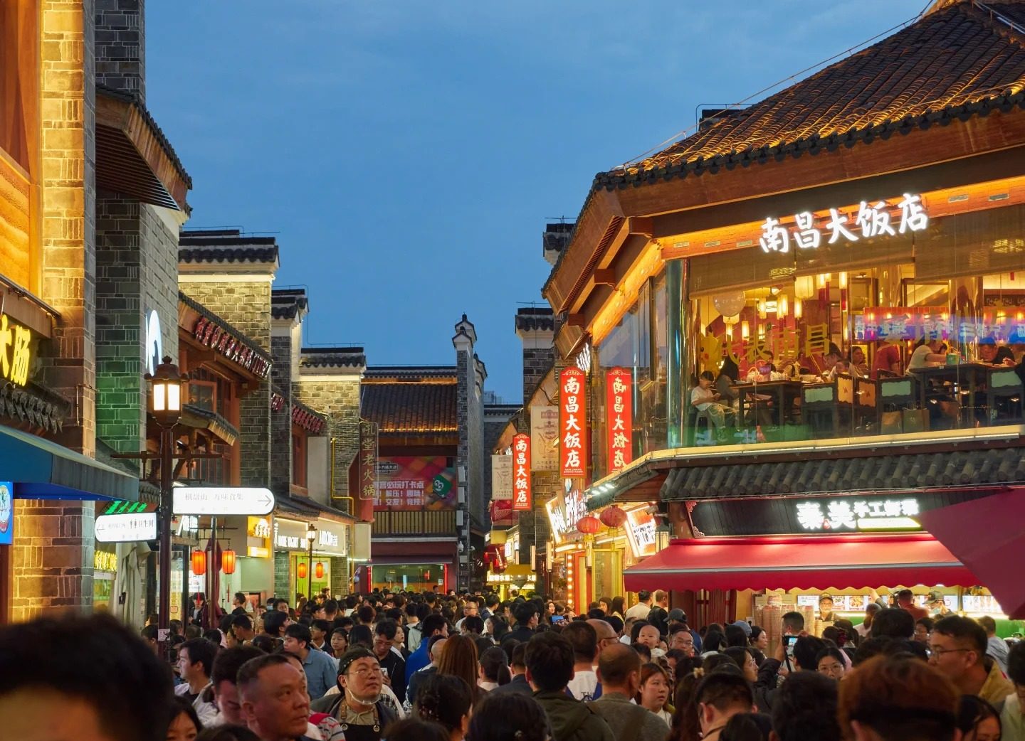 A crowded lane in Nanchang's Wanshou Palace district at blue hour, packed shoulder to shoulder with people beneath lit restaurant signs, red lanterns and grey-tiled roofs