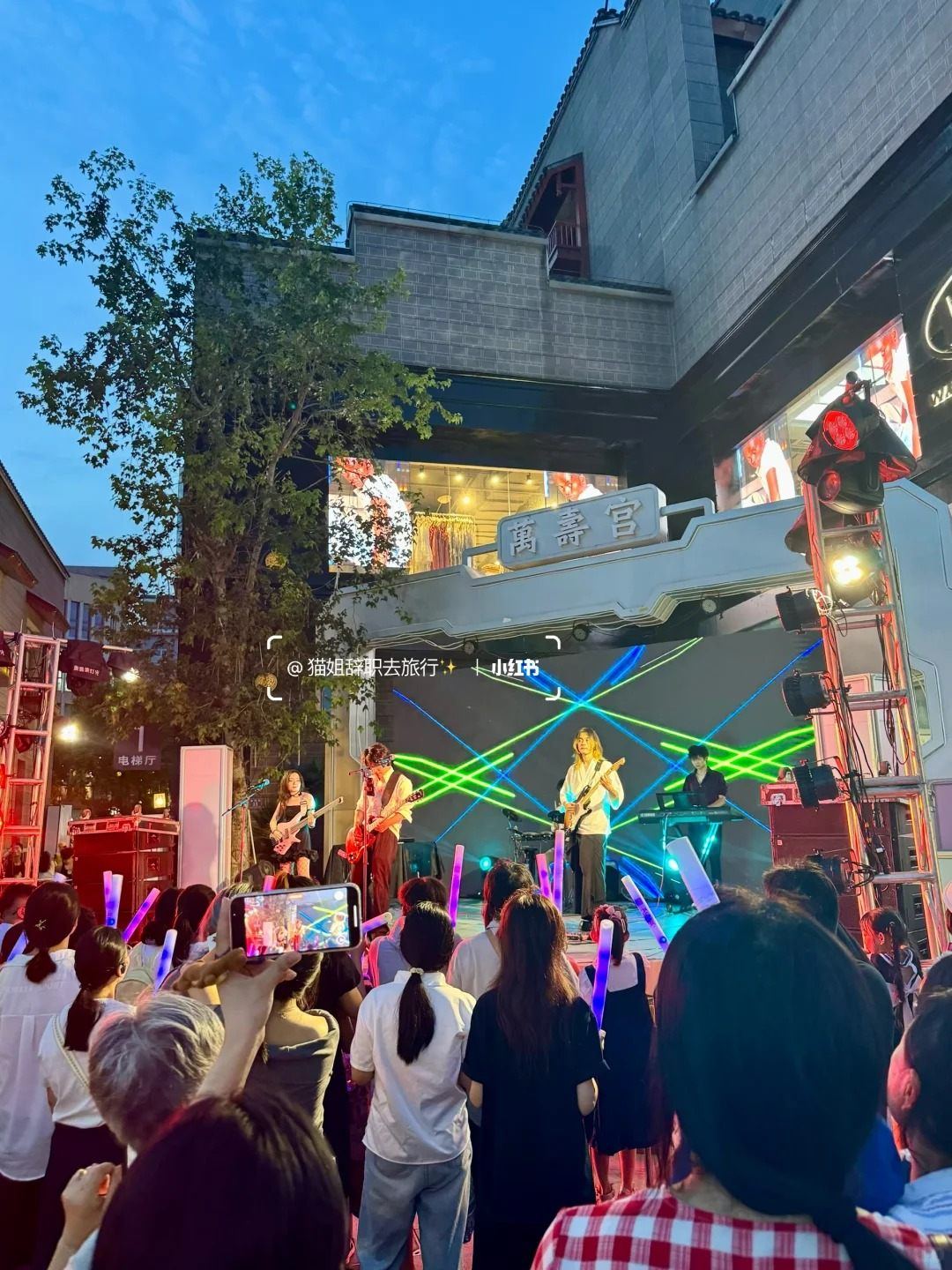 A band playing on an outdoor stage beneath the Wanshou Palace gate in Nanchang, with a crowd holding glow sticks at dusk