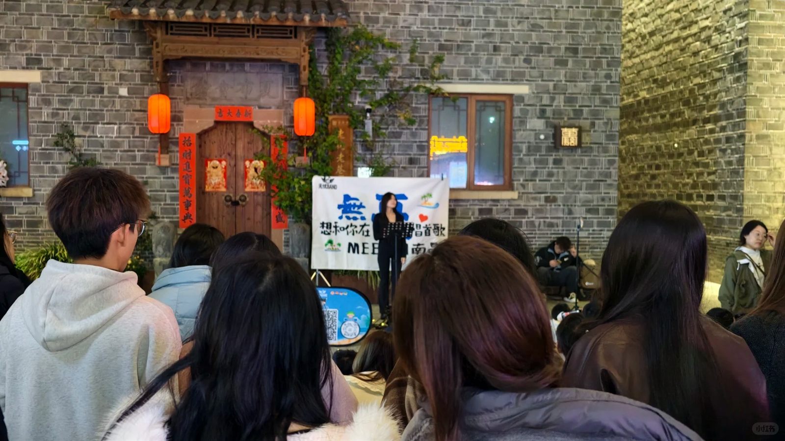 A live singer performs before a seated evening crowd in a lantern-hung courtyard at Wanshou Palace, a grey-brick Gan-style wall behind the small stage