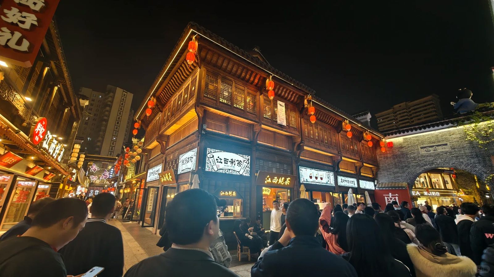 A two-storey timber-latticed Gan-style building in Wanshou Palace at night, grey brick and carved eaves hung with red lanterns above a tea shop, crowds passing below
