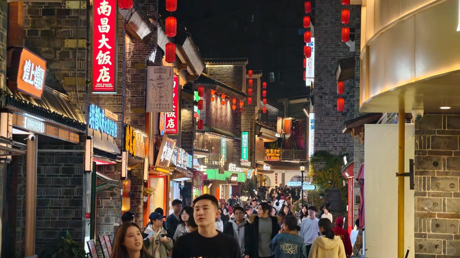 A packed food lane in Wanshou Palace at night, red lanterns and glowing restaurant signboards overhead, crowds shuffling past snack stalls and eateries